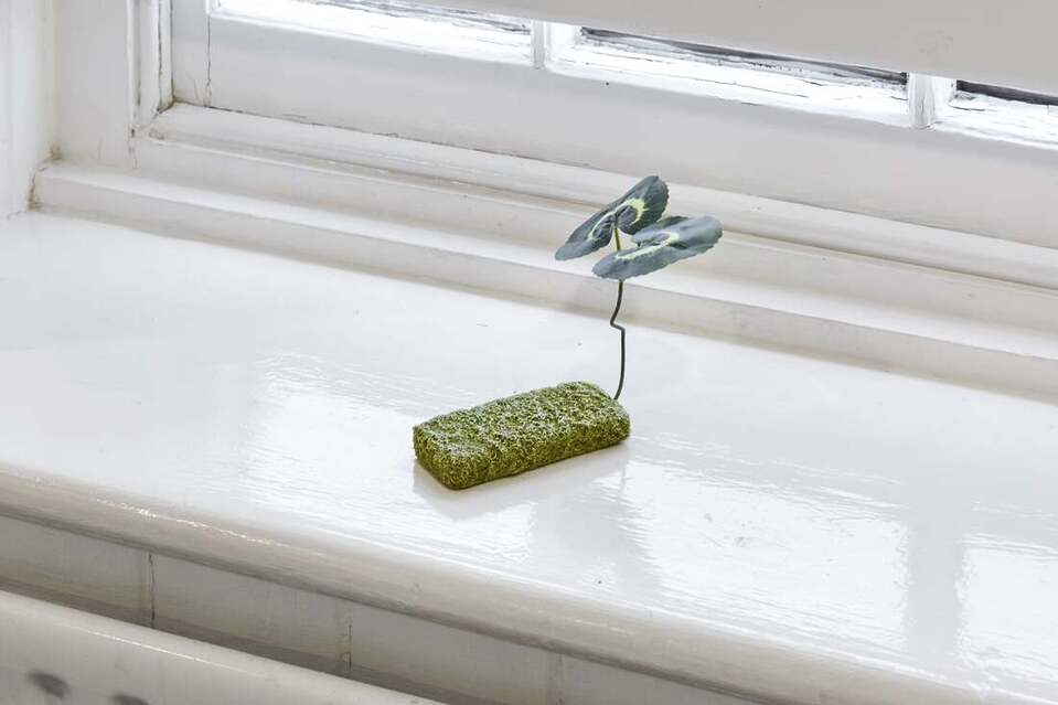 A photo of a white windowsill with a small sculpture placed on it. The sculpture is the shape of an old mobile phone, covered in green moss like texture with a fake plant with two leaves sprouting upwards.